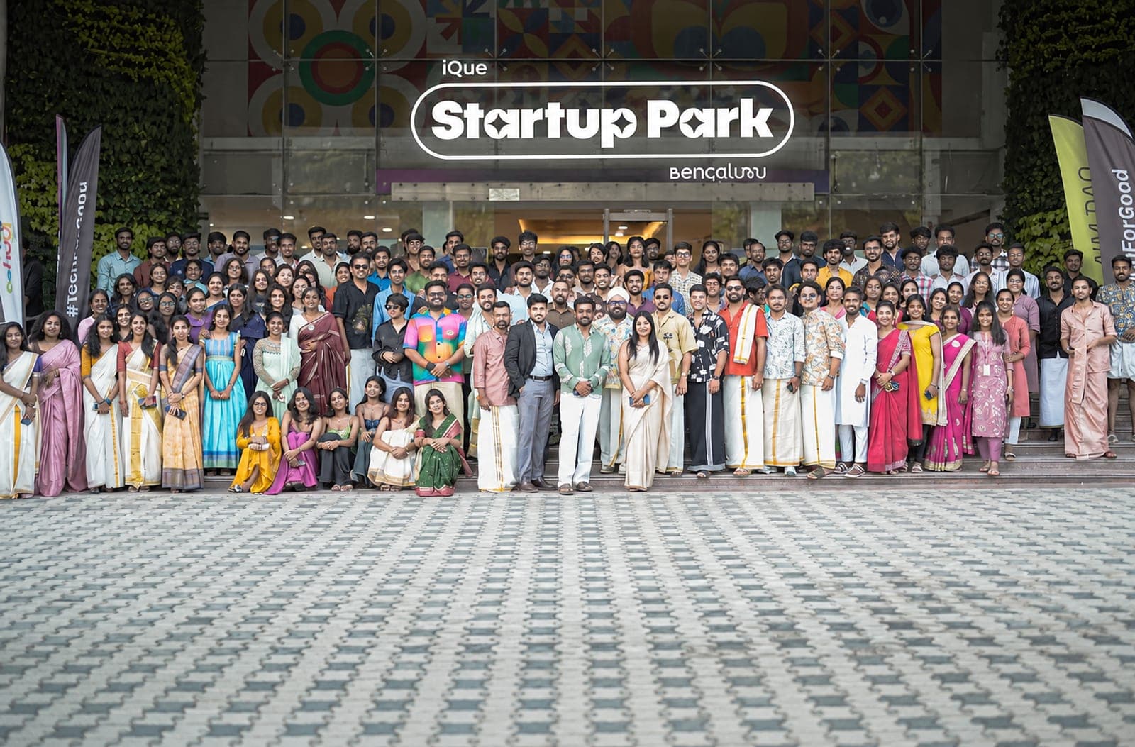 Around two hundred people gathered on the steps of iQue Startup Park Bengaluru beneath the Startup Park sign, many in Kerala festival dress