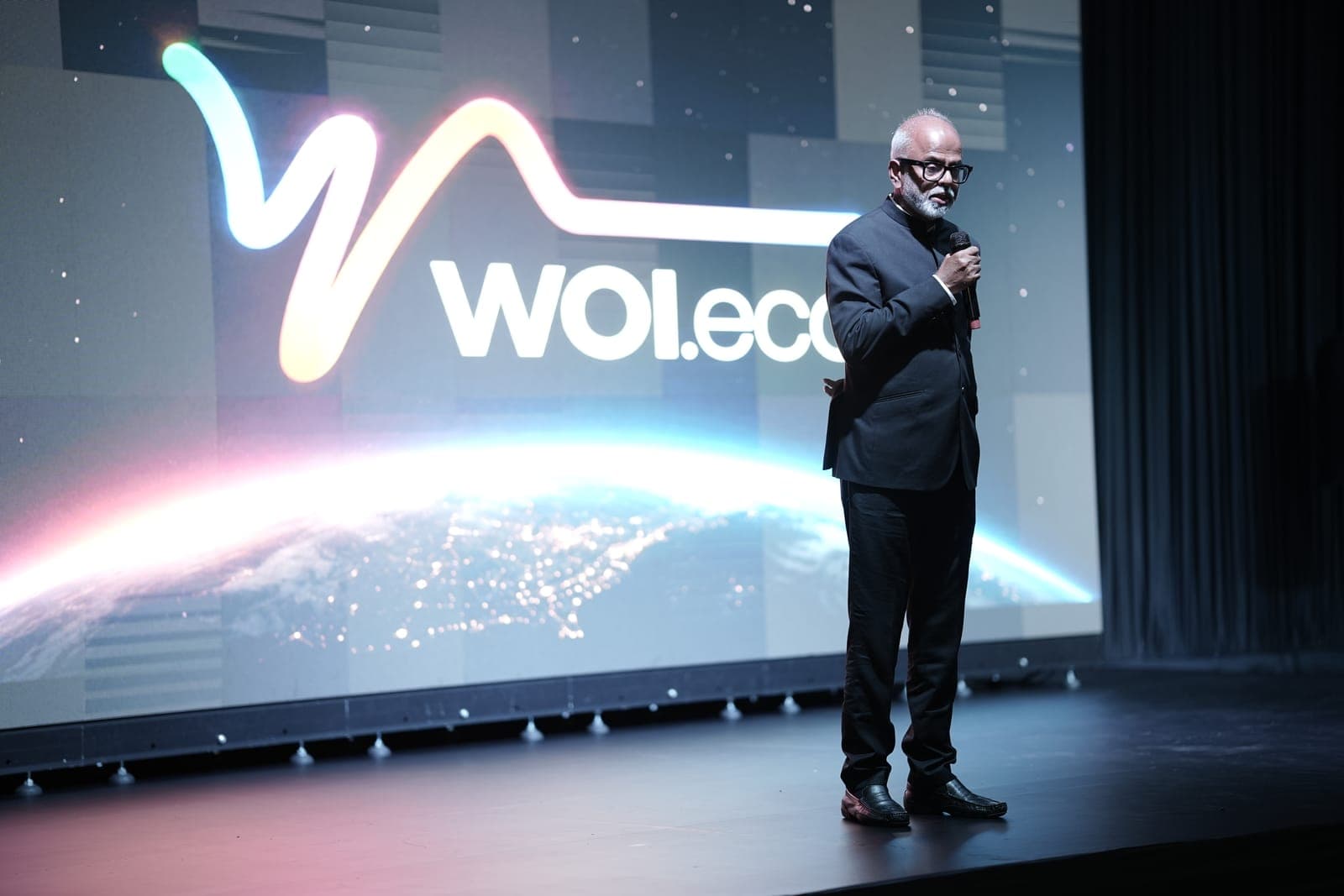 A speaker addressing the room from the stage in front of a large illuminated WOI.eco screen